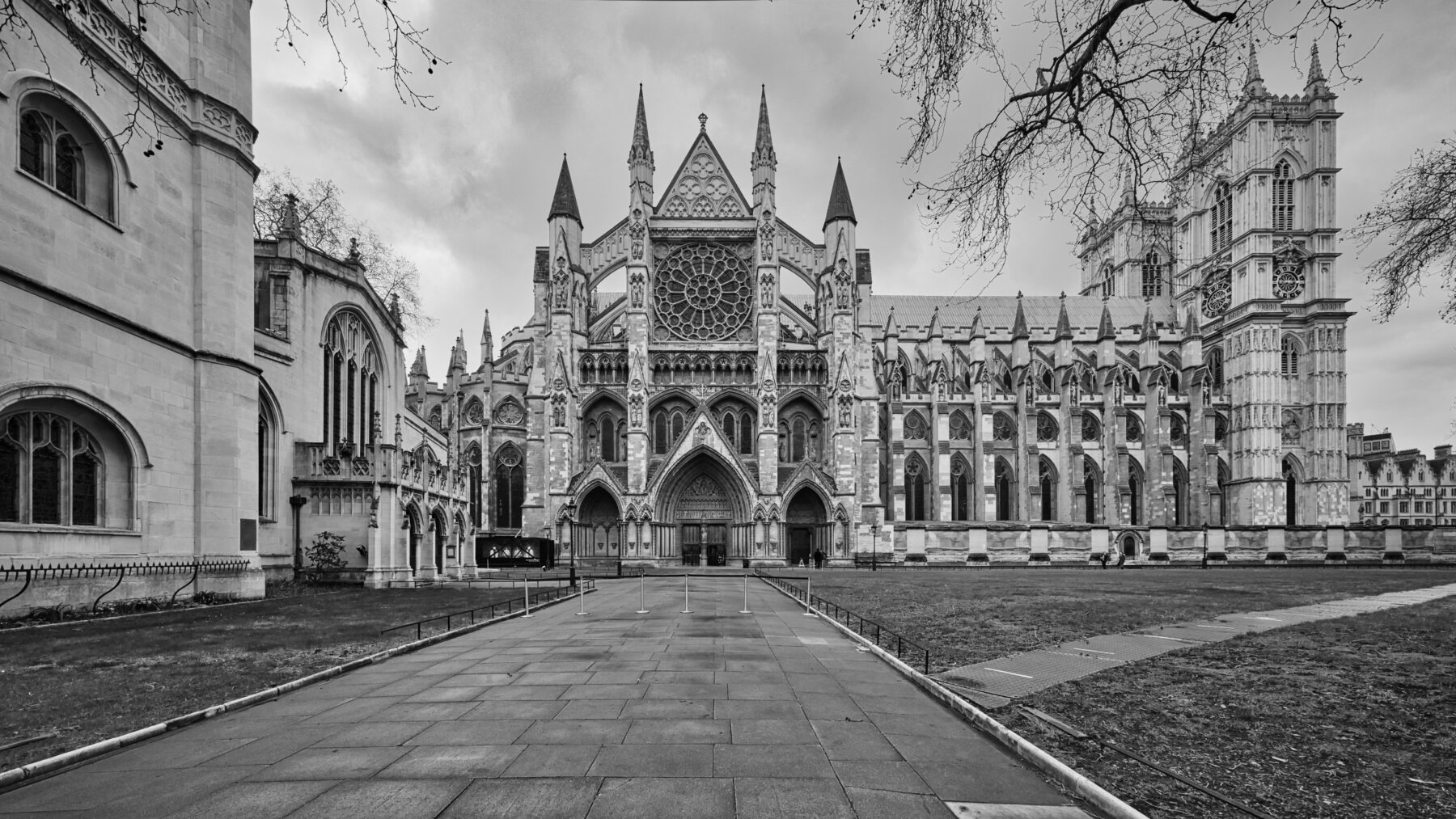 Black-and-white view of the north side of Westminster Abbey across a lawn and paved path under a cloudy sky.