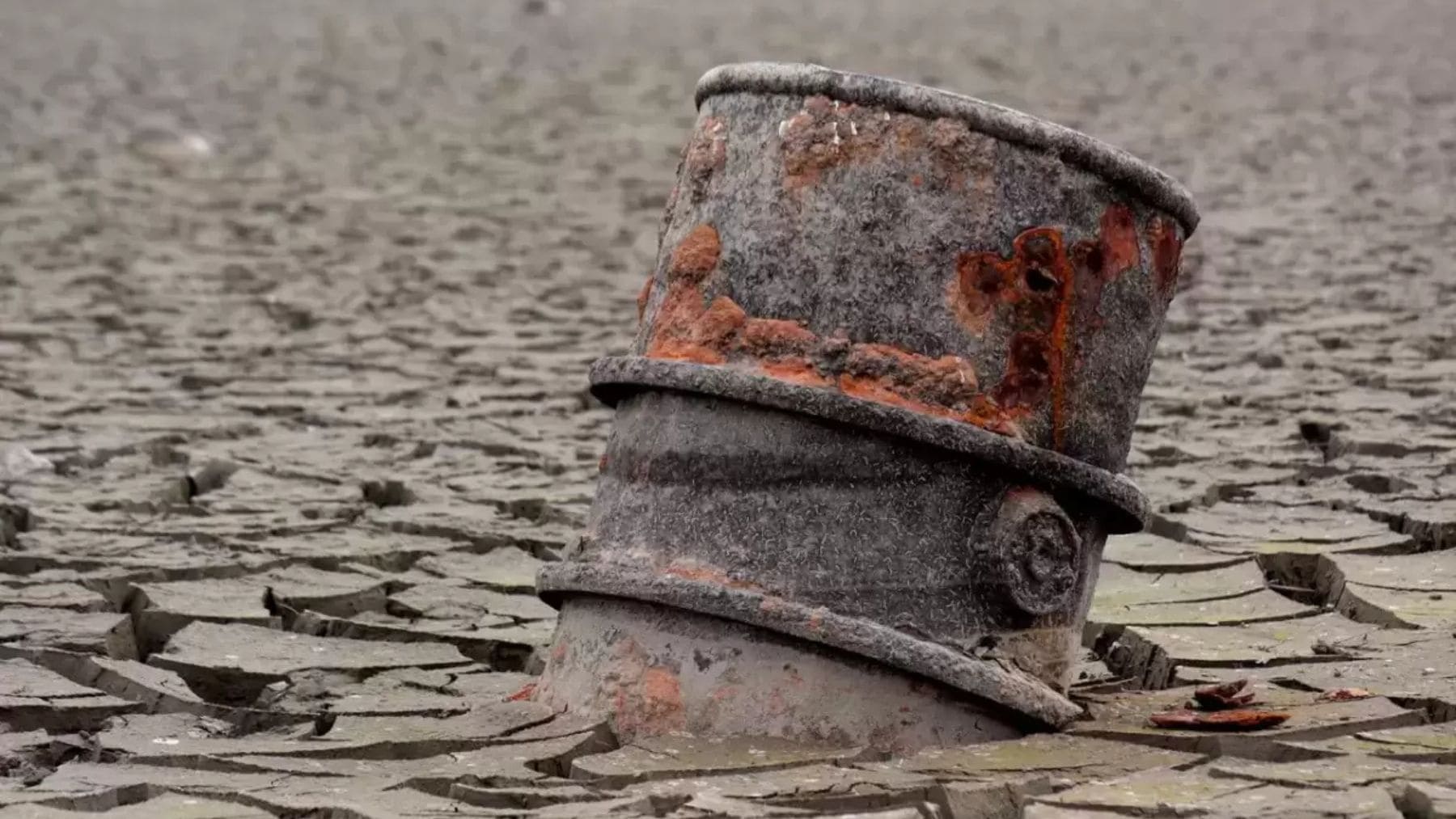 Bidón metálico oxidado semienterrado en suelo agrietado por la sequía, con barro seco y grietas alrededor.