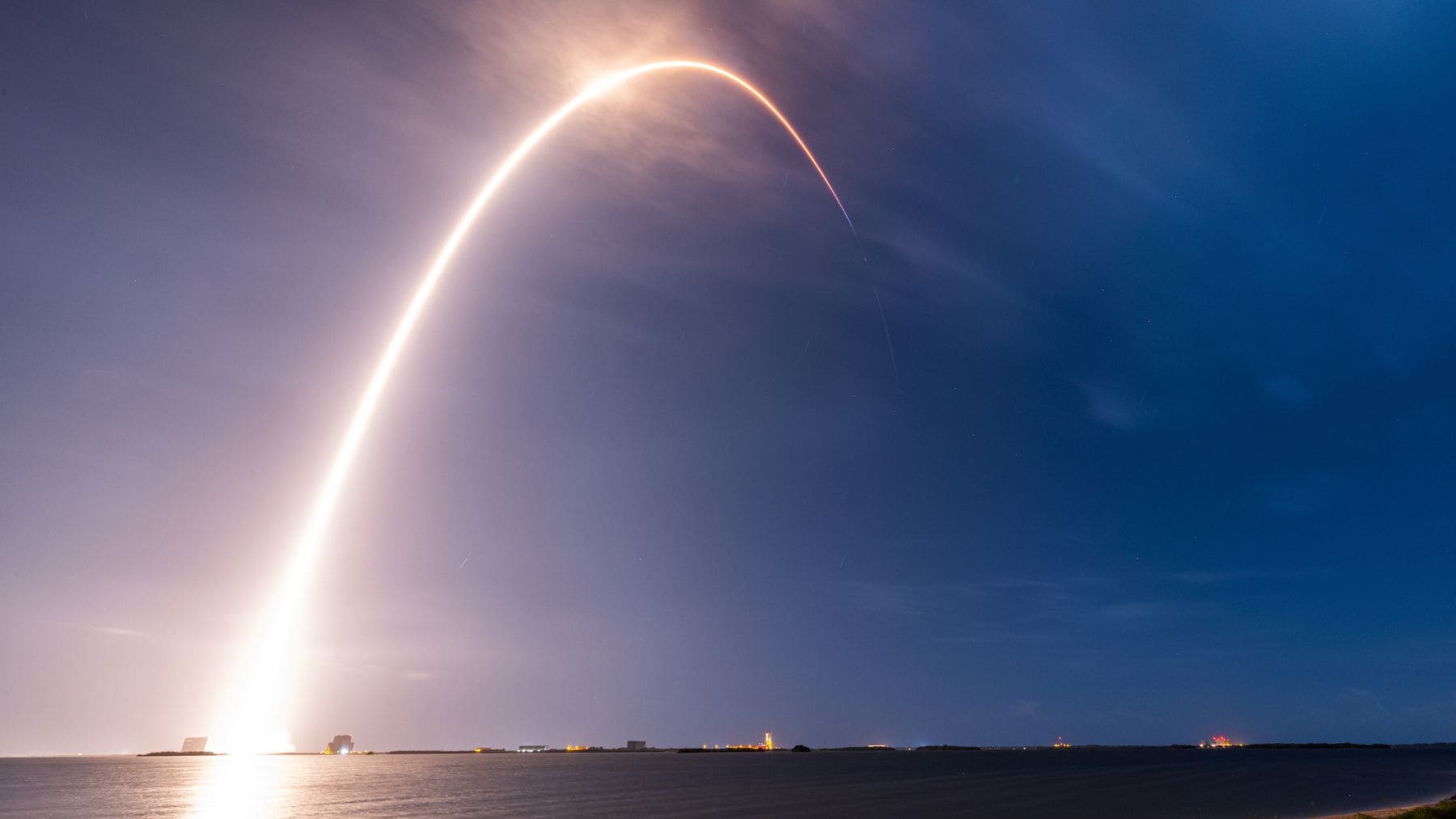 Lanzamiento nocturno de un cohete con estela luminosa en arco sobre el cielo, visto desde la costa junto al mar.