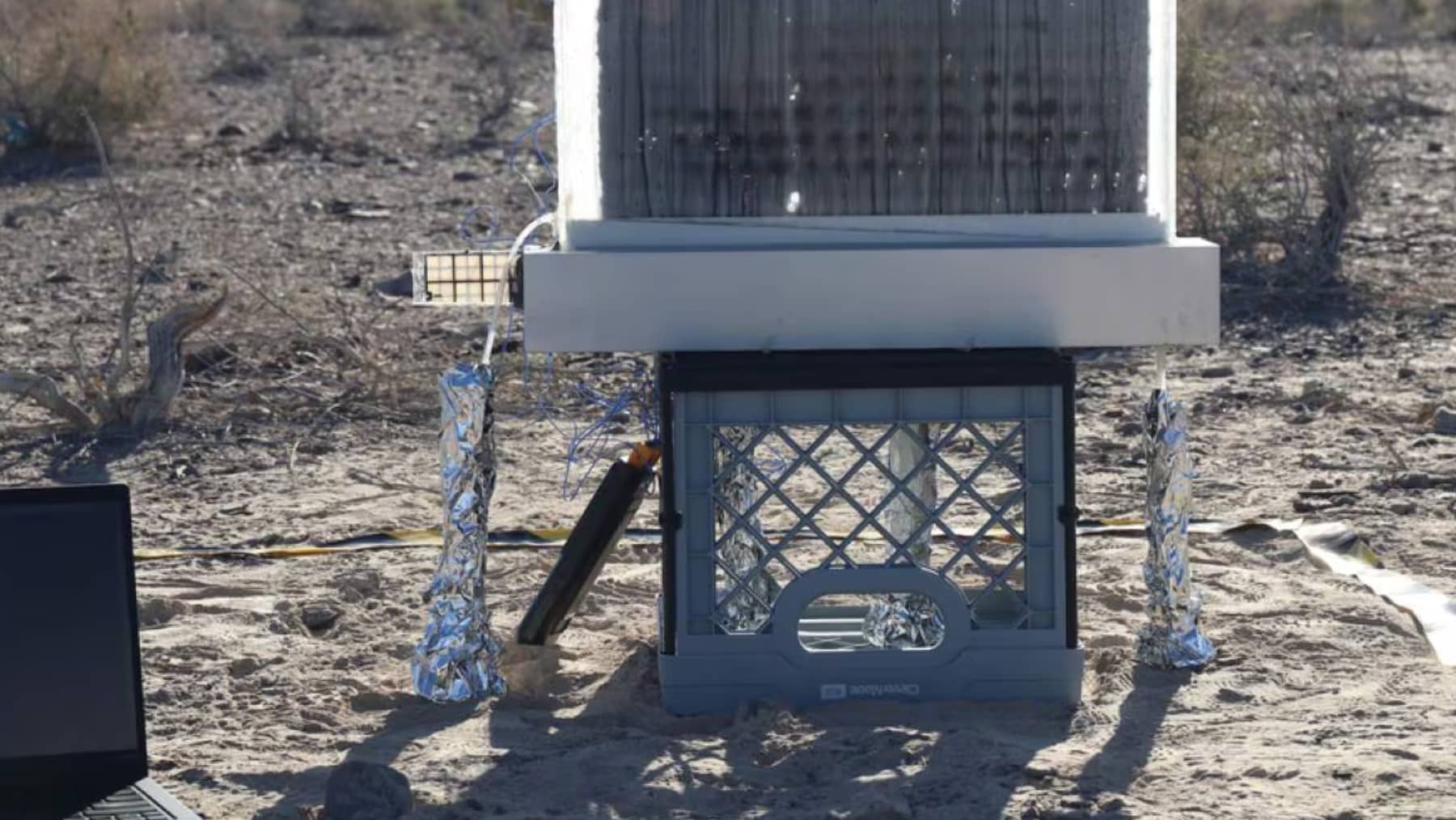 Montaje experimental al aire libre sobre arena con un panel negro en un marco, soportes envueltos en papel de aluminio y una base de caja de plástico.