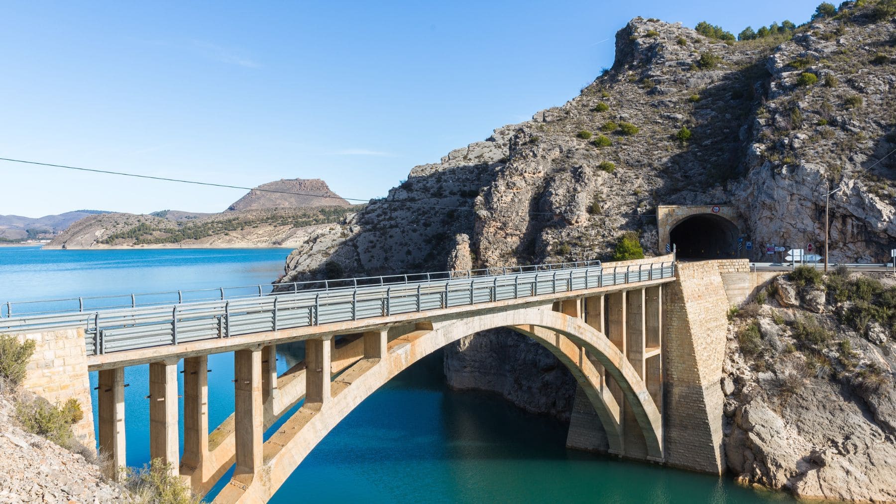 Puente sobre el embalse de La Tranquera en Zaragoza con el agua en tono turquesa y montañas rocosas al fondo.