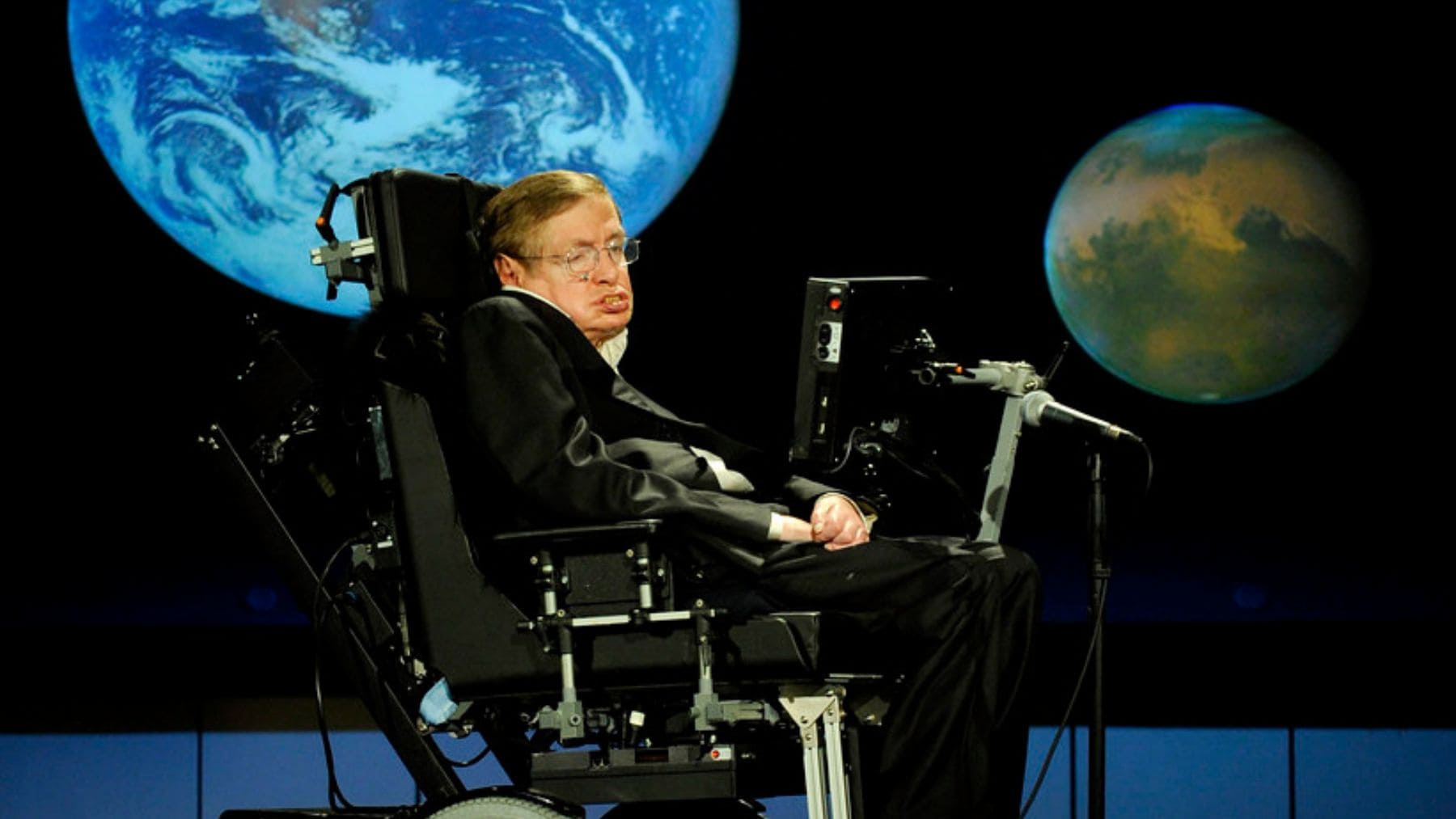 Stephen Hawking durante una conferencia con imágenes de planetas y la Tierra al fondo.