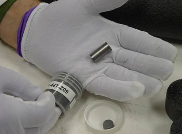 Una mano con guante blanco sostiene un compacto cilíndrico de combustible TRISO
