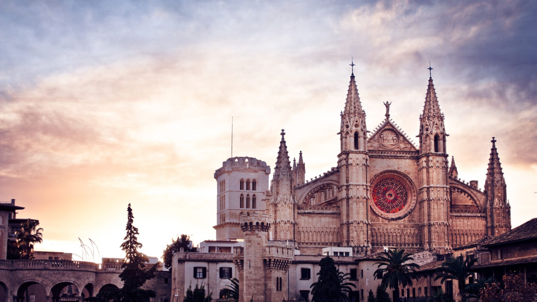 Fachada de la catedral de Palma de Mallorca al amanecer, con el rosetón mayor iluminado de rojo y un cielo rosado y violeta detrás de las agujas góticas