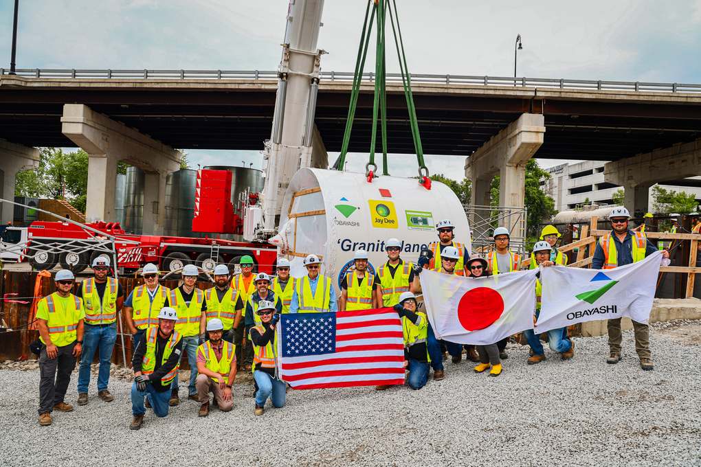Una grúa levanta el escudo de la tuneladora Granite Janet bajo el puente de Queen City Avenue mientras la cuadrilla posa con banderas