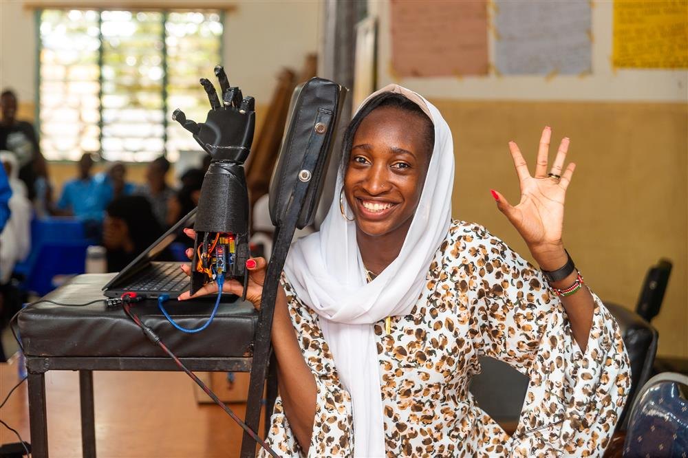 Una joven sonríe junto a una mano robótica negra impresa en 3D con plástico reciclado y su electrónica a la vista