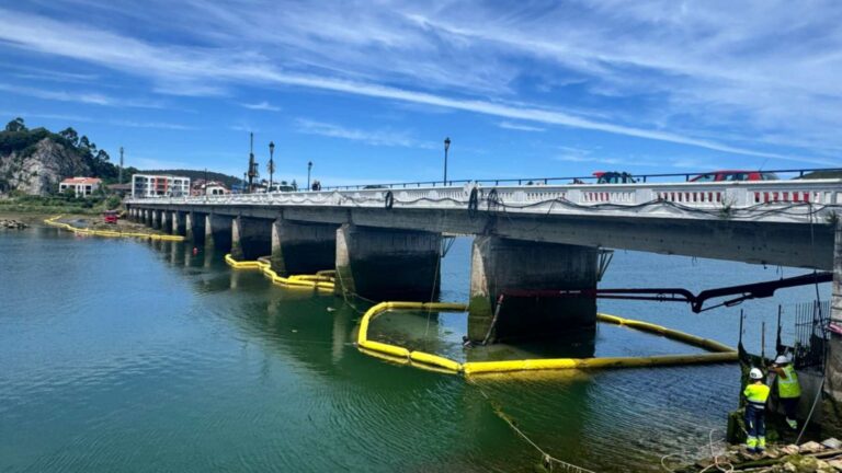 Obras en el puente de Ribadesella sobre el río Sella, con barreras antiturbidez amarillas alrededor de las pilas y dos operarios en la orilla