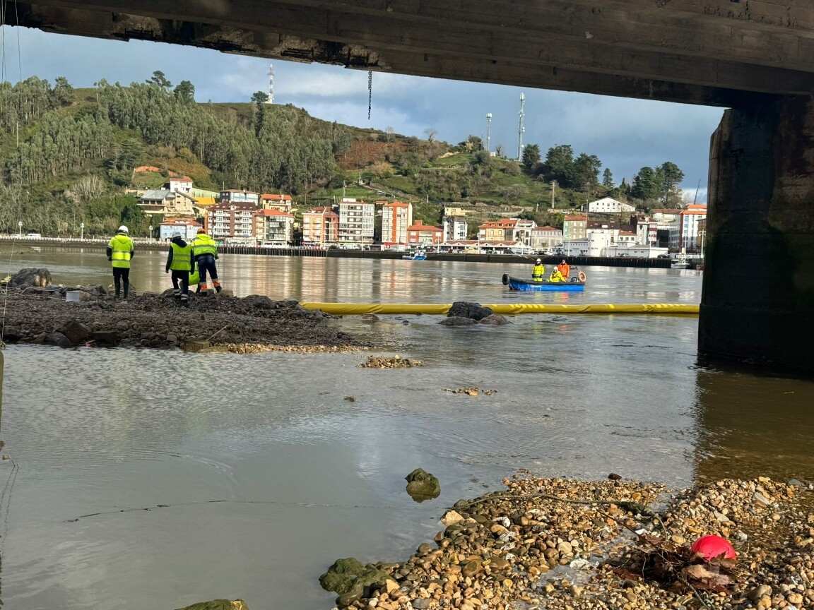 Operarios sobre una escollera bajo el puente de Ribadesella y una embarcación junto a una barrera antiturbidez en la ría del Sella