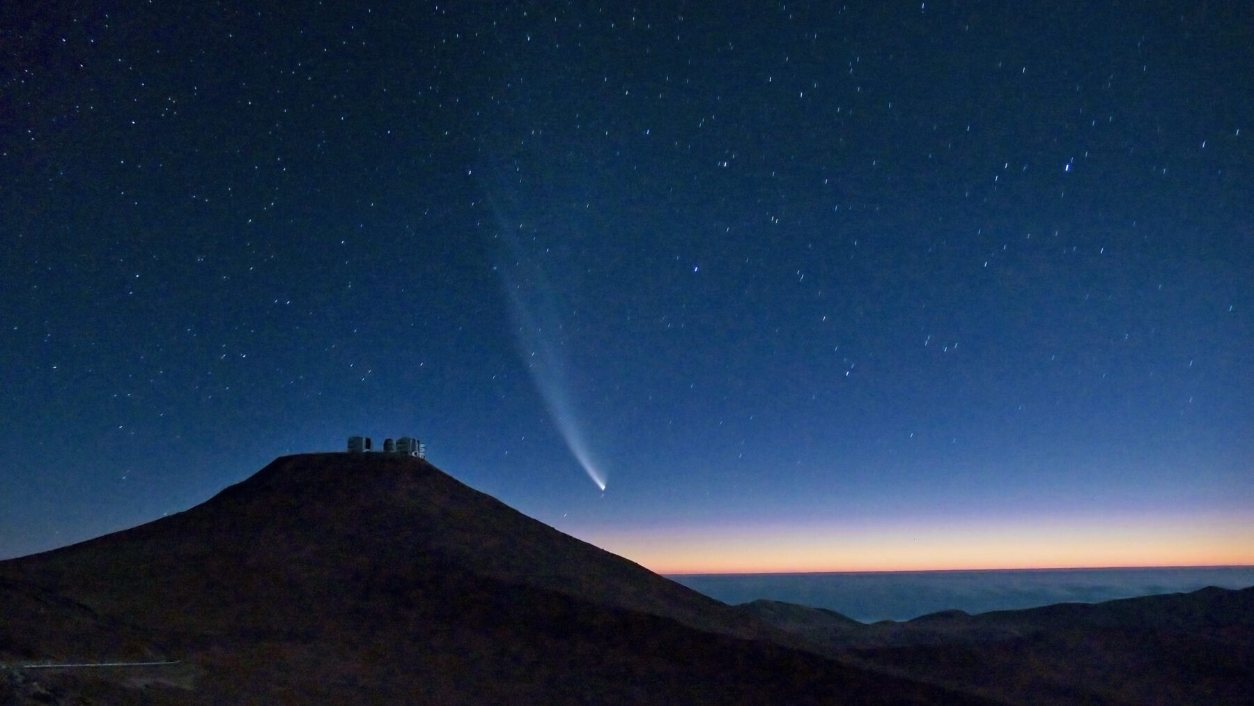 Cometa con una larga cola curva sobre el cerro Paranal, en Chile, en un cielo estrellado al atardecer