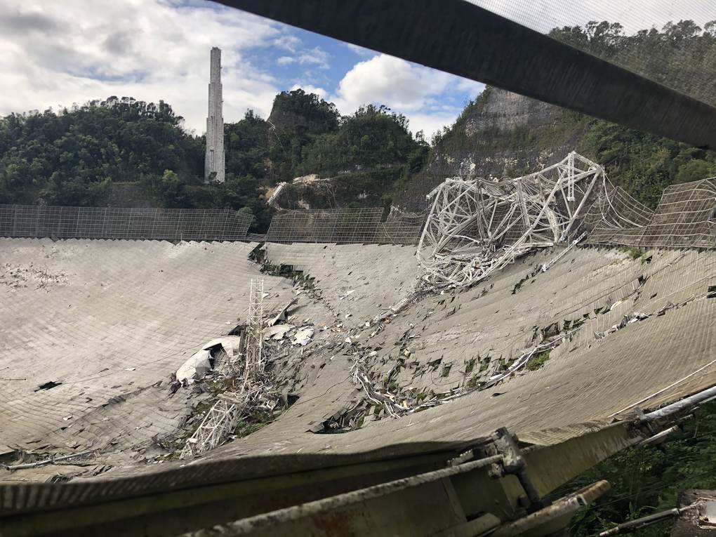 Reflector del observatorio de Arecibo con paneles rotos y la estructura de la plataforma caída sobre el plato, con una de las torres al fondo