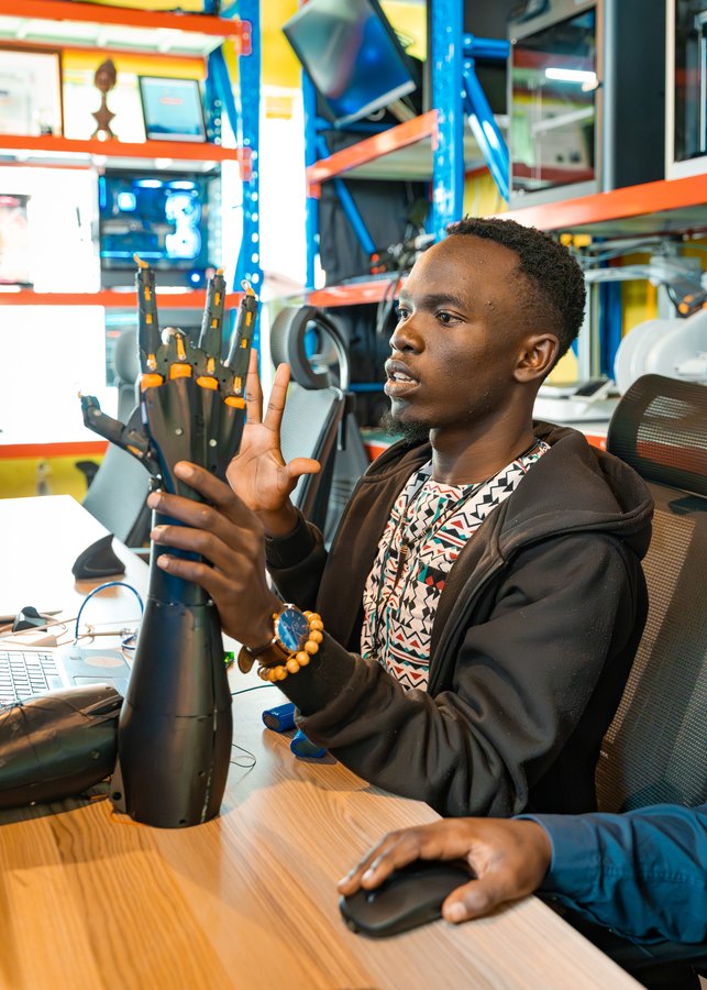 Un joven hace un signo con la mano junto a una mano robótica impresa en 3D en el taller de ZeroBionic