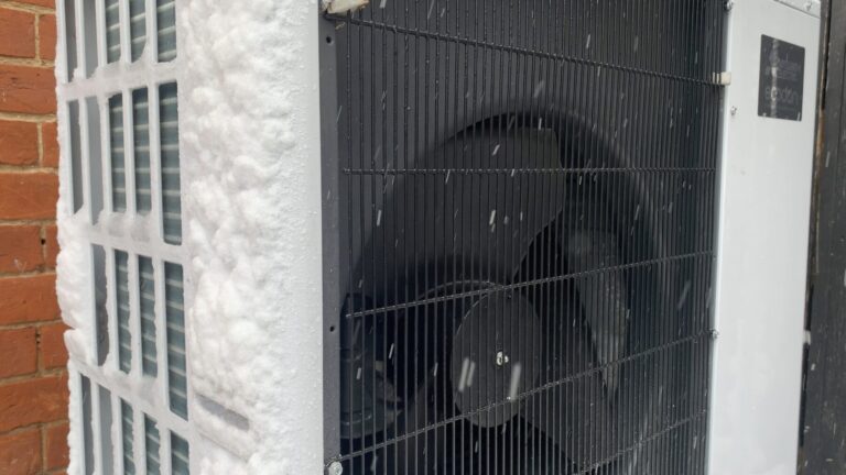 Unidad exterior de una bomba de calor aire-agua cubierta de escarcha y nieve, con copos cayendo delante de la rejilla del ventilador y un muro de ladrillo a la izquierda