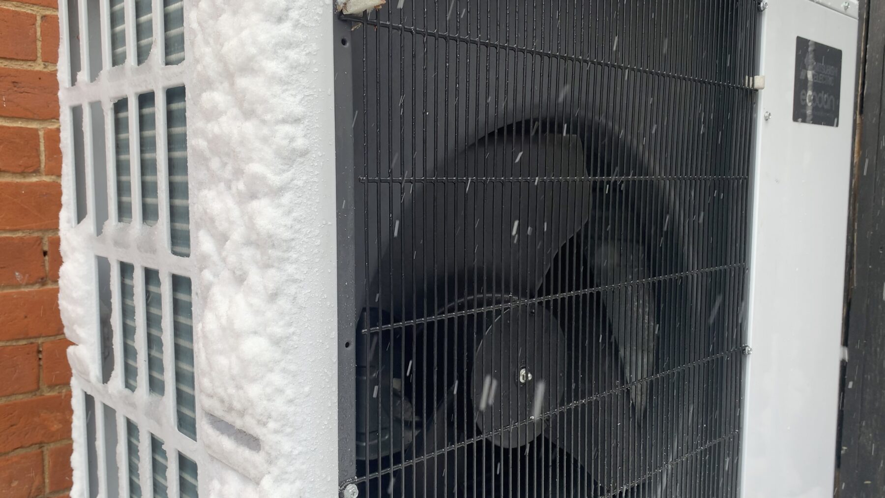 Unidad exterior de una bomba de calor aire-agua cubierta de escarcha y nieve, con copos cayendo delante de la rejilla del ventilador y un muro de ladrillo a la izquierda