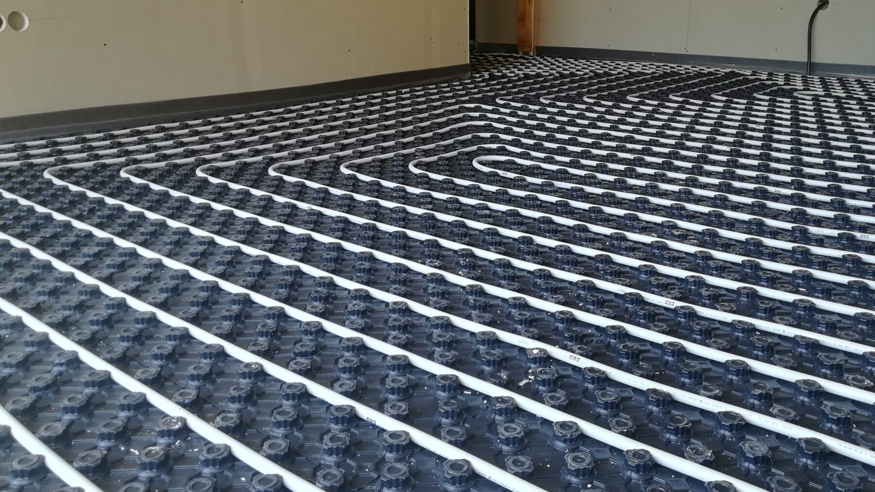 Tuberías blancas de suelo radiante dispuestas en serpentín sobre una placa de nódulos oscura, en el suelo de una vivienda en construcción