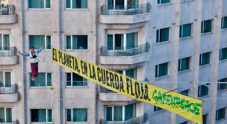 Activista de Greenpeace sobre una cinta de highline a 30 metros de altura en el centro de Madrid, junto a una gran pancarta con el mensaje “El planeta, en la cuerda floja”, como protesta ante la COP30 de Belém