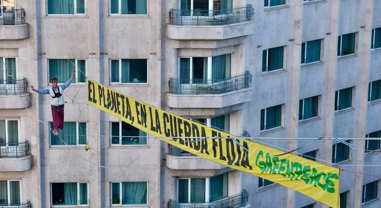 Activista de Greenpeace sobre una cinta de highline a 30 metros de altura en el centro de Madrid, junto a una gran pancarta con el mensaje “El planeta, en la cuerda floja”, como protesta ante la COP30 de Belém