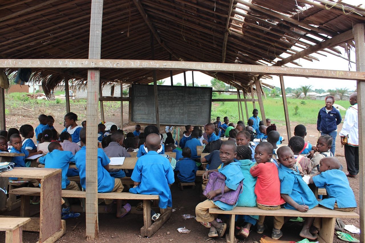 Niños y niñas de Sudán del Sur asistiendo a clase en una escuela sencilla con techo de chapa y ventilación, rodeada de terreno inundado o muy seco, simbolizando la educación y la resiliencia climática frente a las inundaciones y sequías