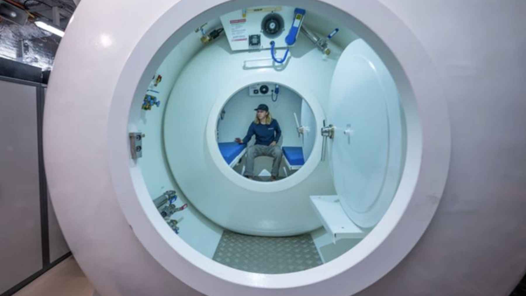 Paciente sentado dentro de una cámara hiperbárica de oxigenoterapia con puerta circular abierta.