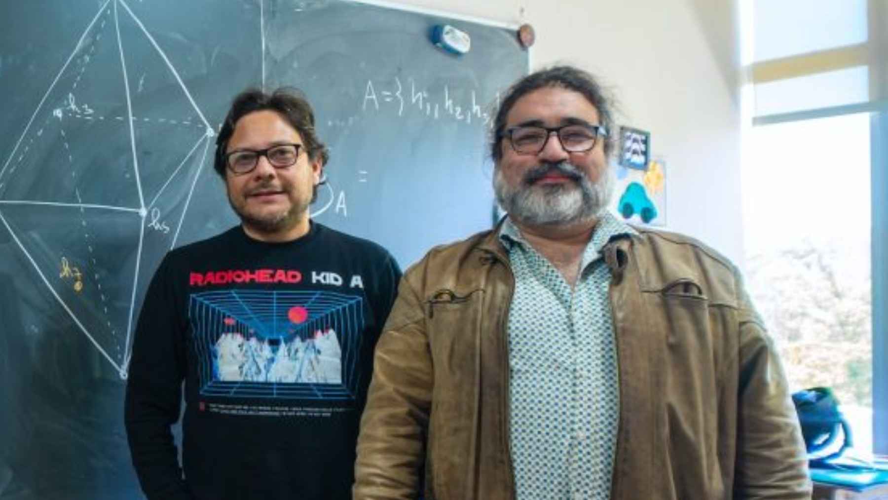 Dos investigadores posan delante de una pizarra con un esquema geométrico en un aula