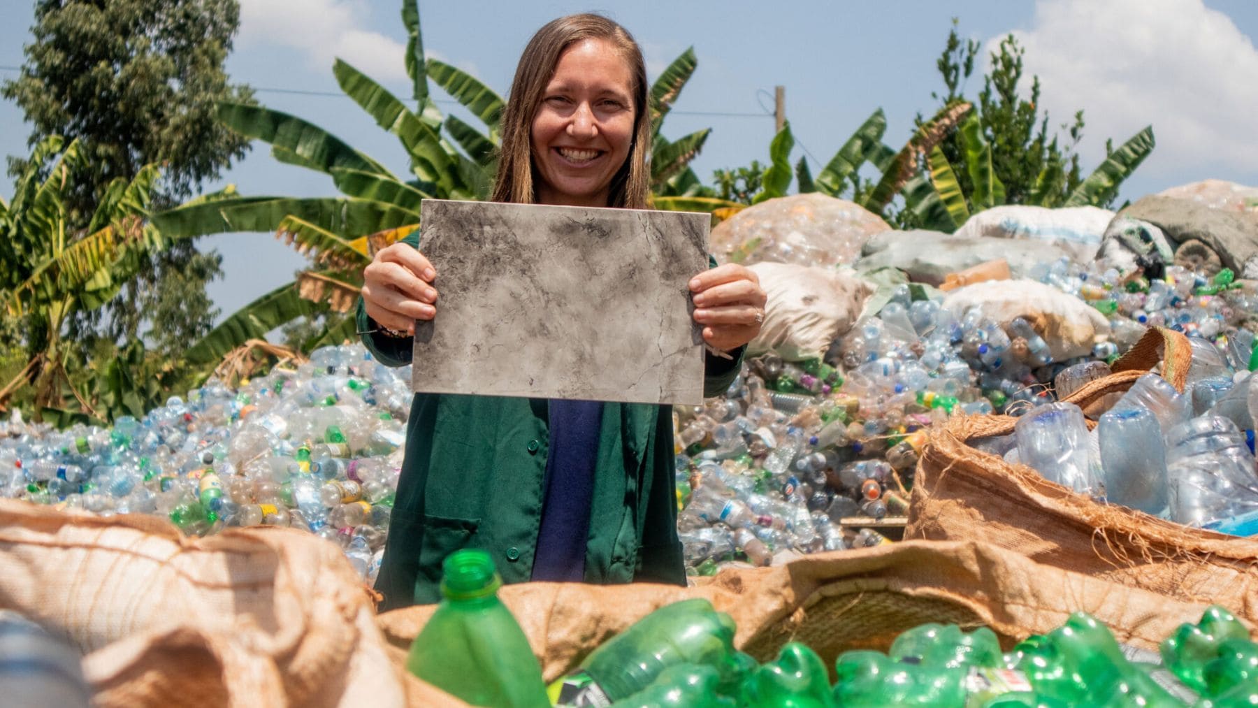 Emprendedora en Uganda muestra baldosas hechas con plástico reciclado entre montañas de botellas PET.