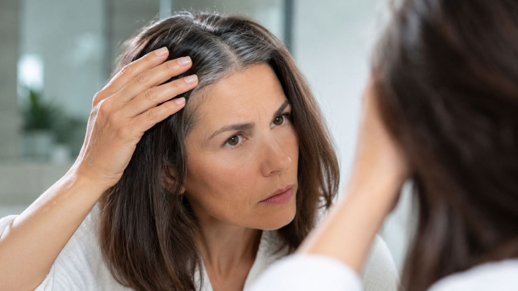 Mujer observándose las primeras canas frente al espejo tras un estudio que investiga cómo revertir el encanecimiento del cabello.