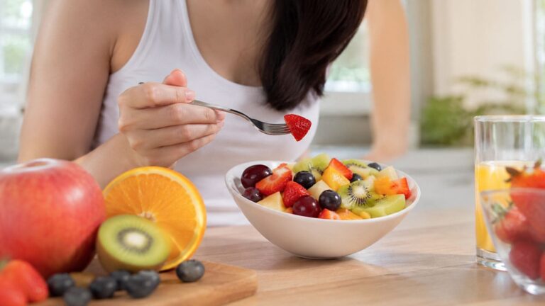 Mujer comiendo un bol de fruta fresca con fresas, kiwi, naranja y uvas durante el desayuno en una cocina luminosa.