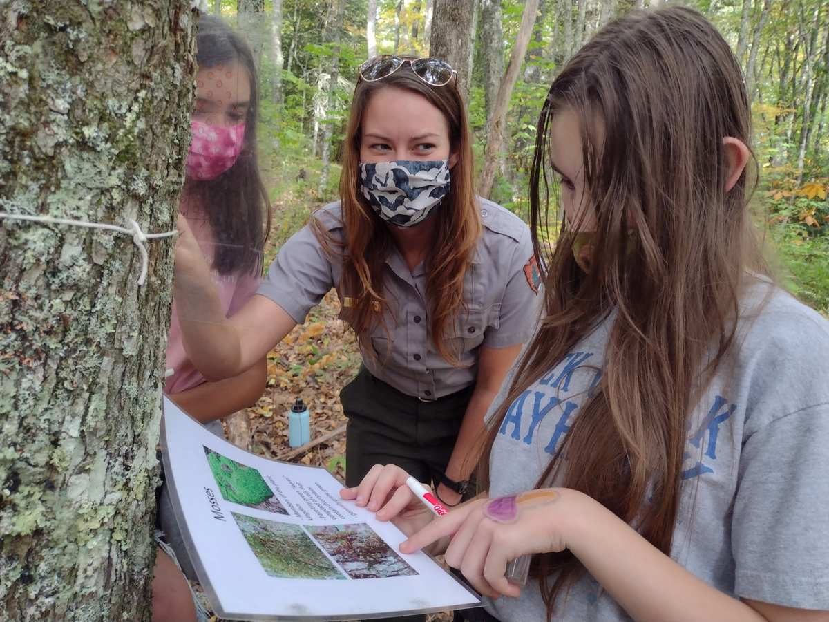 Una guarda con uniforme y dos alumnas con mascarilla miran una ficha junto a un árbol