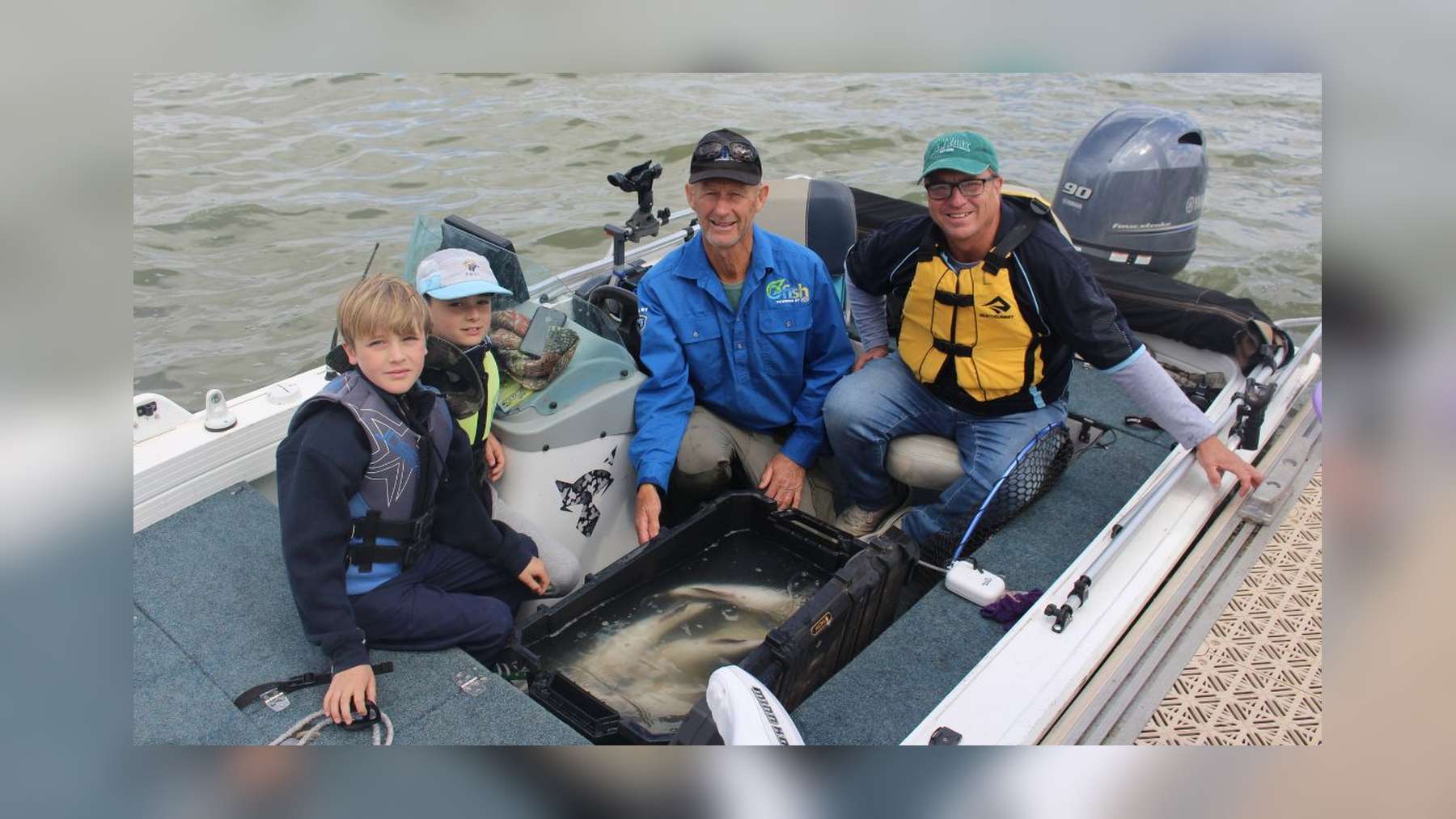 Dos adultos y dos niños en una barca del lago Bonney junto a una cubeta con bacalaos de Murray antes de la suelta